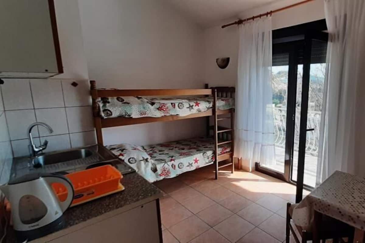 Une petite cuisine équipée d’un évier, d’une bouilloire et d’une table avec une chaise. On aperçoit un lit superposé à l’arrière-plan.