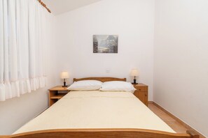 Un lit simple avec un cadre en bois, deux tables de chevet avec des lampes, un tableau accroché au mur et une fenêtre avec des rideaux blancs.