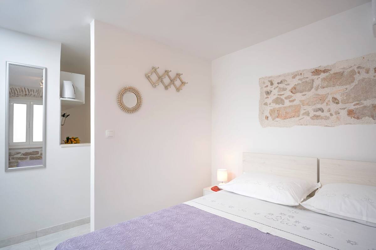 A bedroom with a bed, a nightstand, a wall-mounted mirror, a stone wall accent, and a shelf with a white pot.