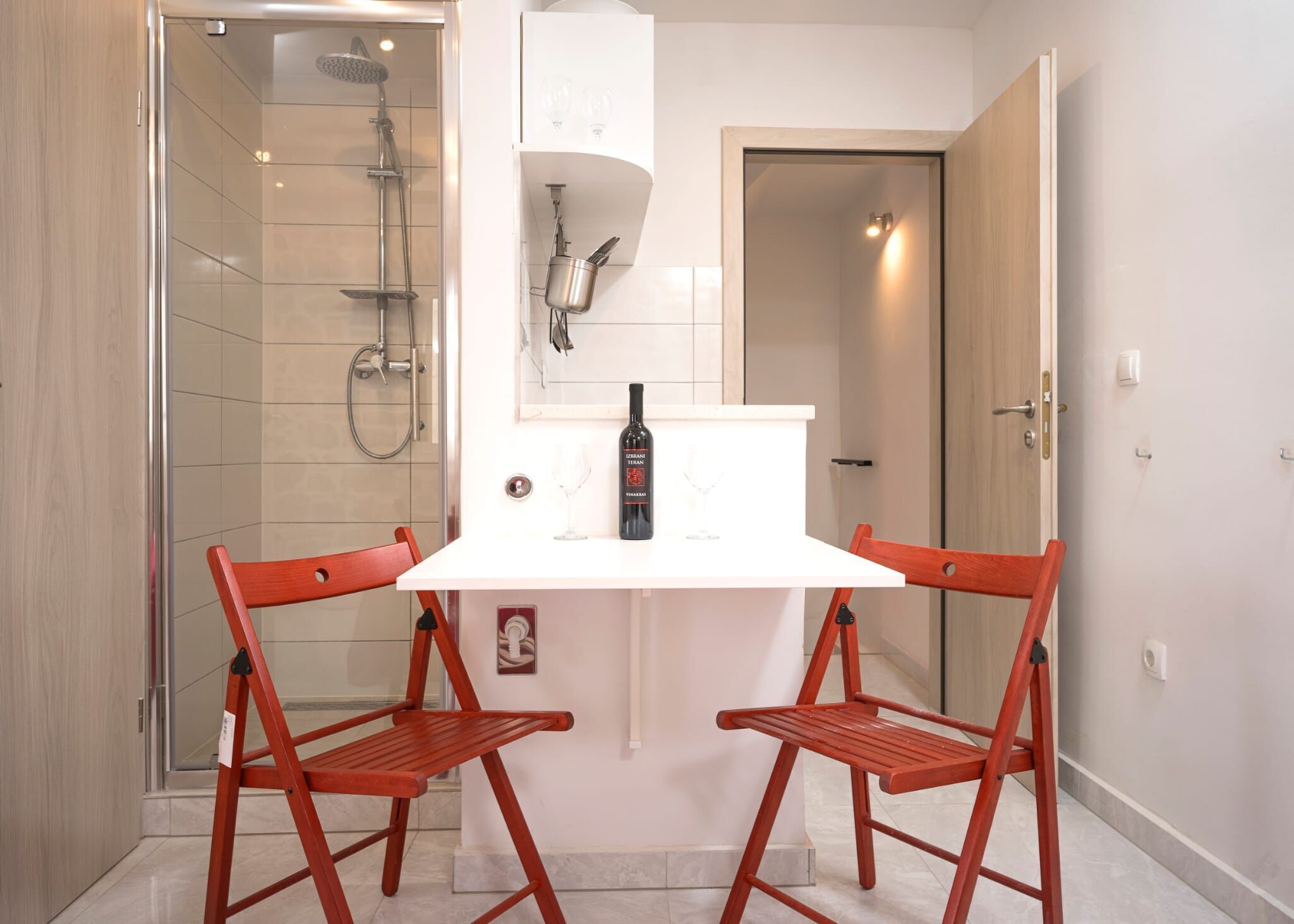 Habitación pequeña con ducha, una mesa blanca y dos sillas rojas, además de una botella de vino sobre la mesa.