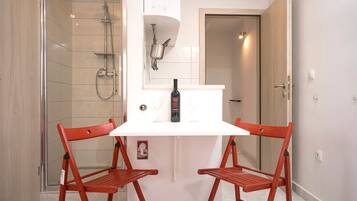 Habitación pequeña con ducha, una mesa blanca y dos sillas rojas, además de una botella de vino sobre la mesa.