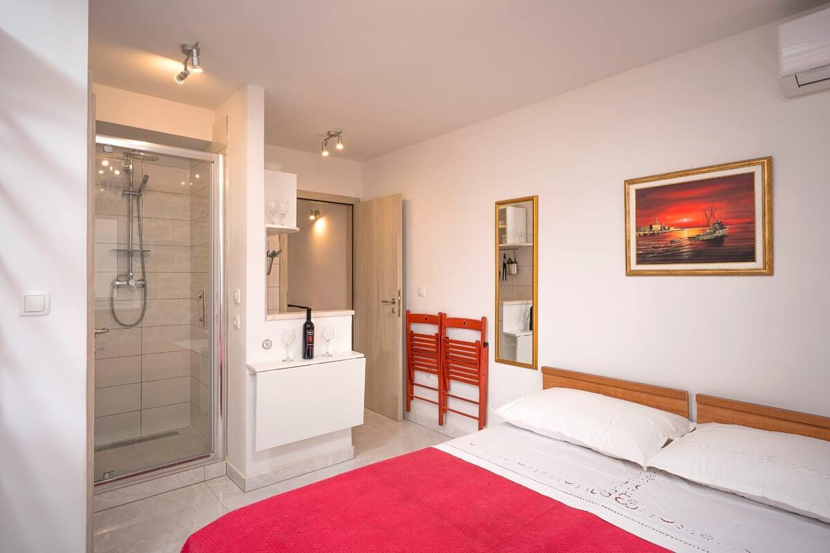 A hotel room with a shower, a wooden chair, a bed with white bedding, a red rug, and a framed picture on the wall.