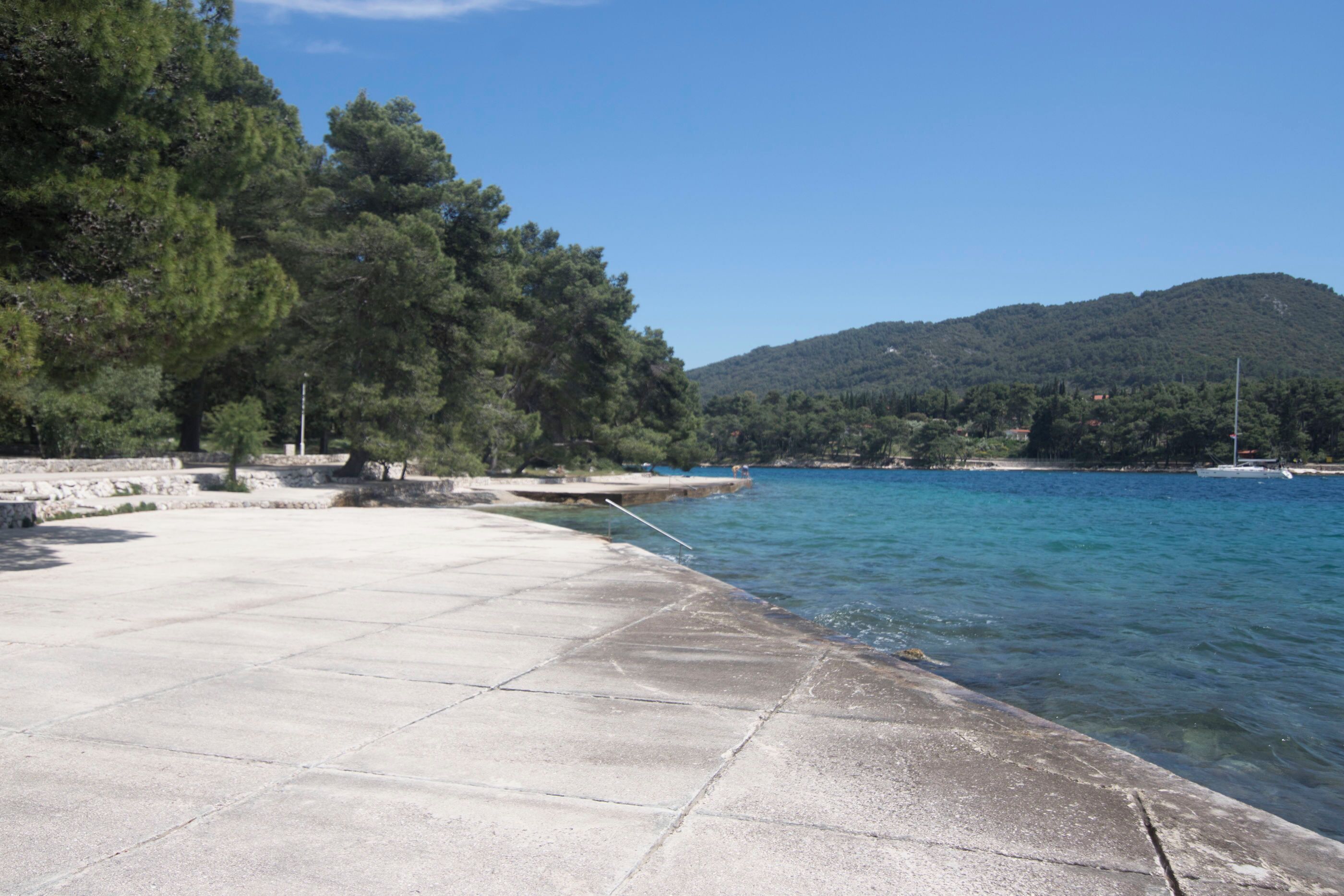 A concrete waterfront promenade with a clear view of the sea and a forested hill in the background.