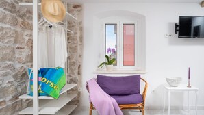 Une pièce avec un mur en pierre, une étagère blanche où sont posées des serviettes, une chaise en osier, une petite table avec une bougie et un téléviseur fixé au mur.