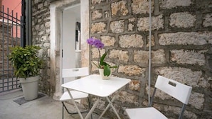 Un petit espace extérieur aménagé avec une table blanche et deux chaises blanches, un pot de fleurs et une orchidée violette.
