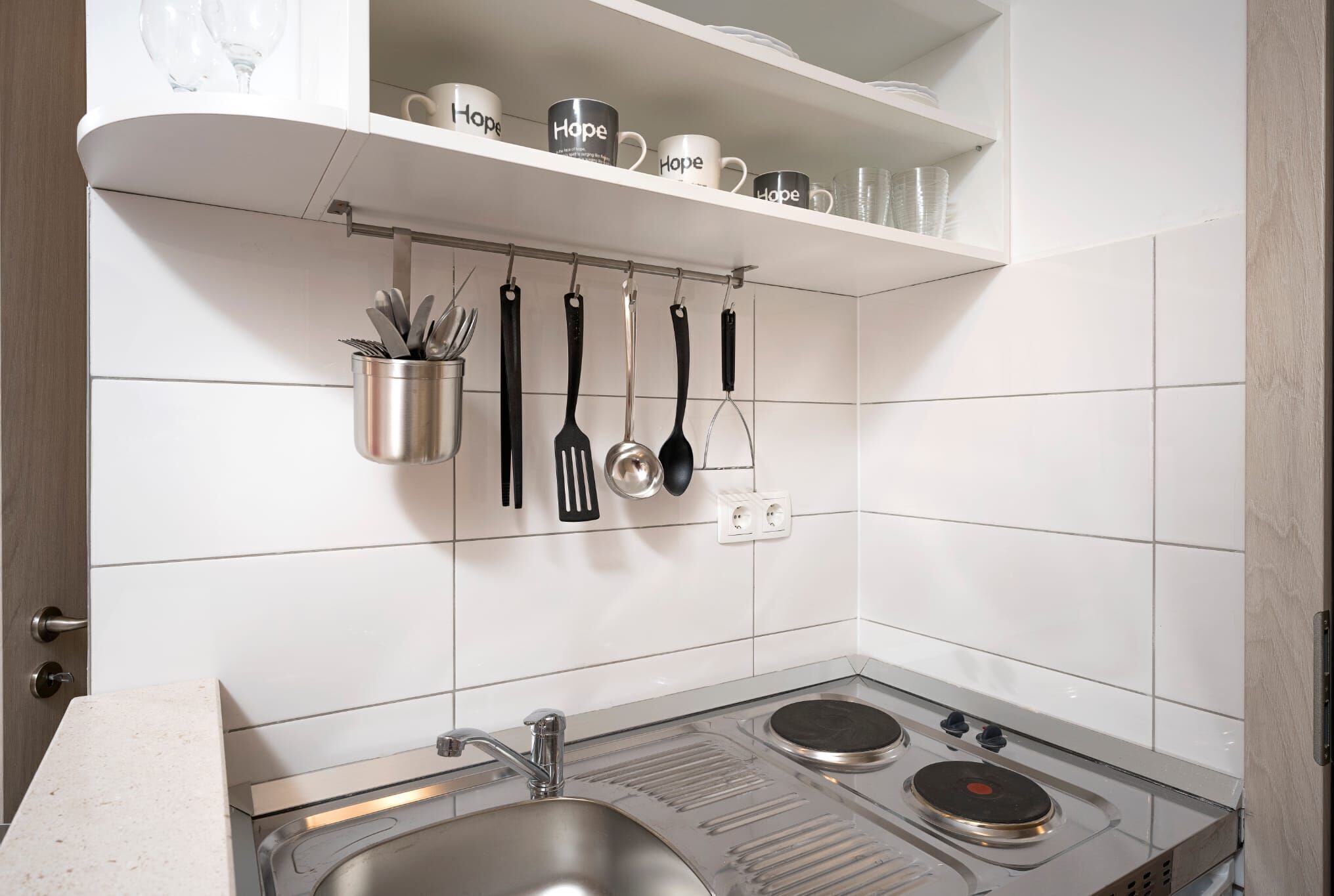 A kitchen with white tiled walls, a stainless steel sink, and a stove. There are utensils hanging on a rack above the sink.