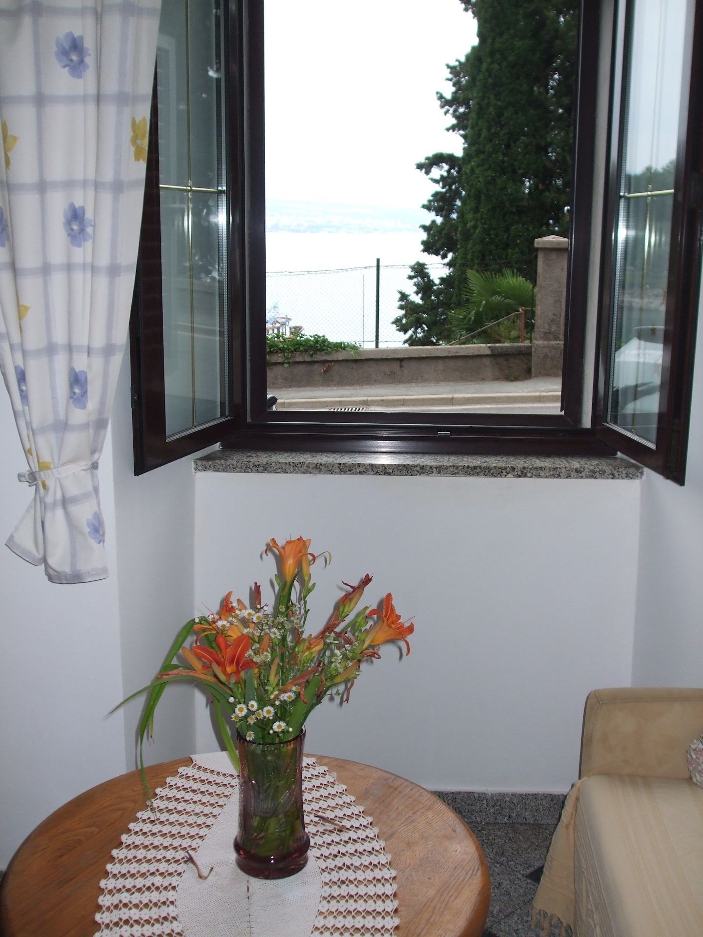 A room with a window overlooking a street, a vase with flowers on a table, and a chair.
