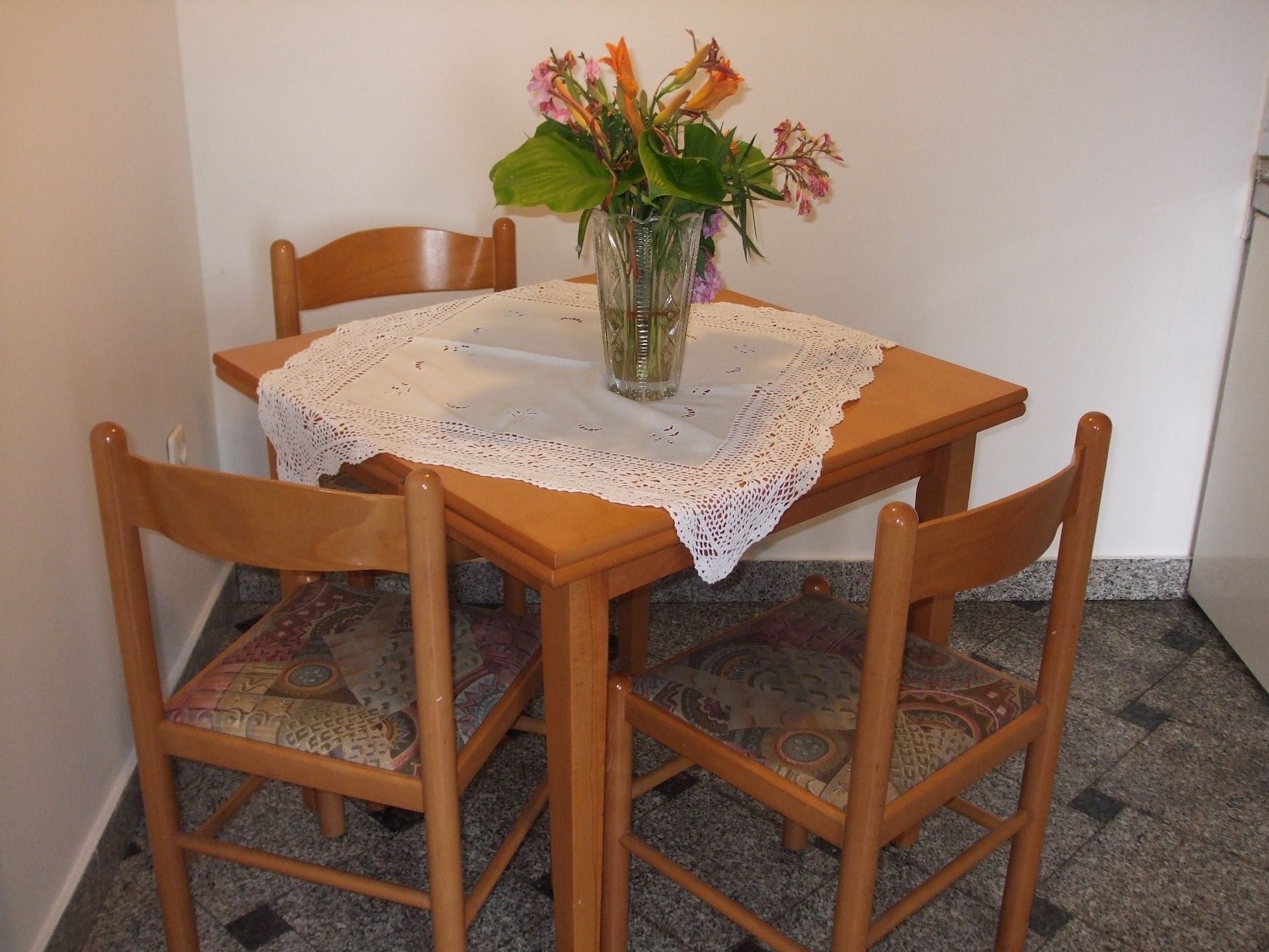 A dining table with a lace tablecloth, surrounded by wooden chairs with patterned cushions.
