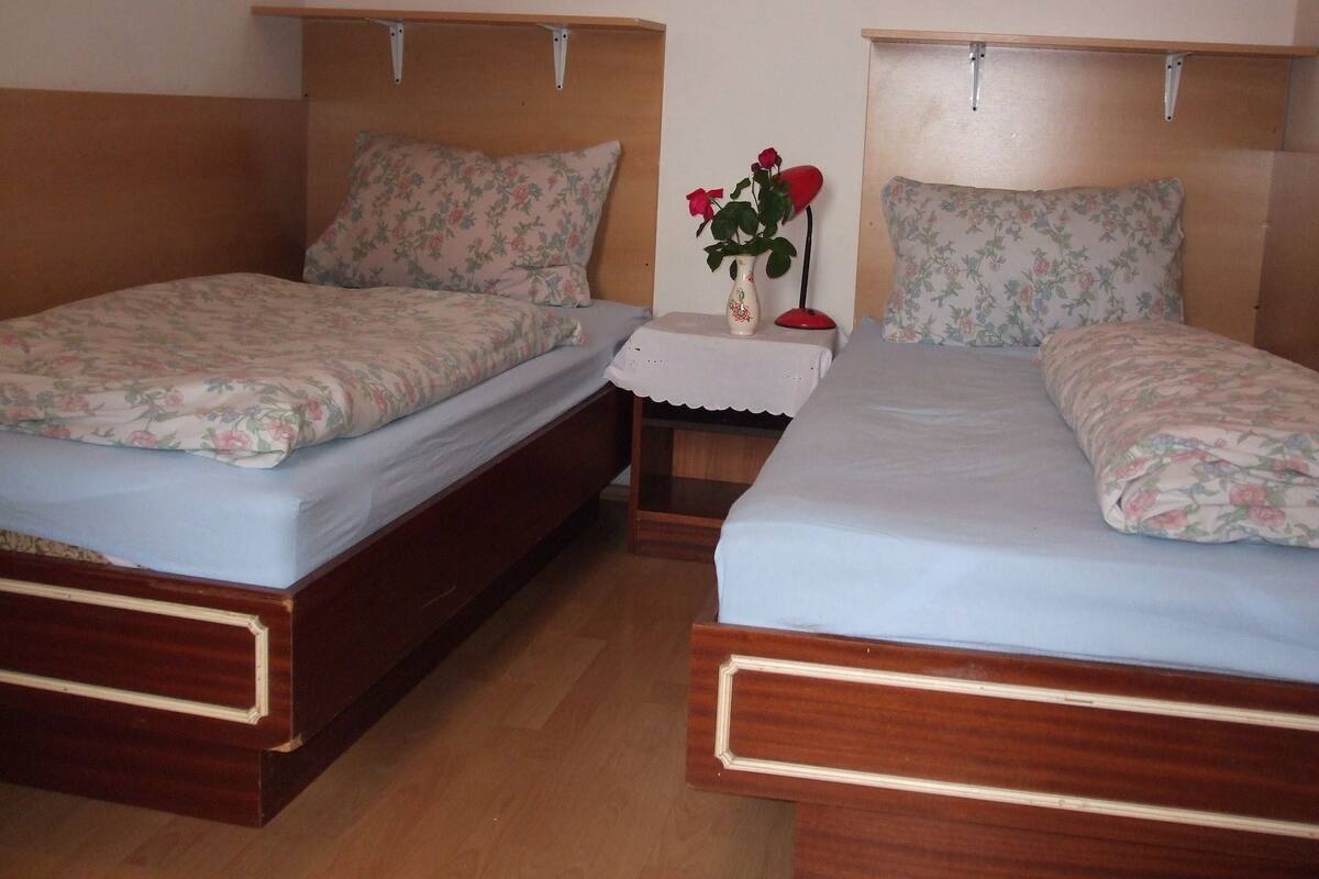 Two wooden beds with floral bedding, a nightstand with a vase of flowers, and a wooden floor.