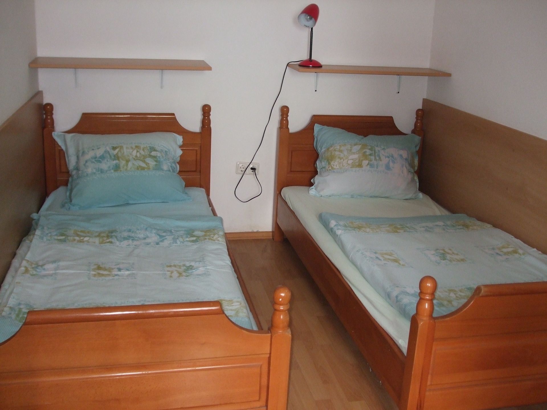Two wooden beds with floral bedding, a red lamp on a shelf, and a power outlet on the wall.