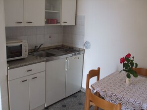 A compact kitchen with a microwave, sink, and a small refrigerator. There's a table with a lace tablecloth and a vase with flowers. - Apartments Vesna (Opatija)