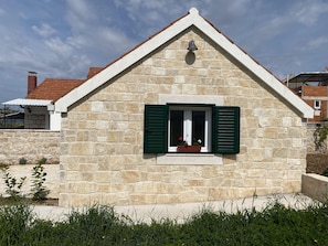 A stone house with a gabled roof, a small window with green shutters, and a bell above the window. - House Little Flower 1 (Okrug)