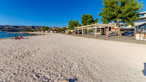 Ein Strand mit Kieselsteinen, einem überdachten Sitzbereich und Blick auf das Meer sowie Gebäude im Hintergrund.