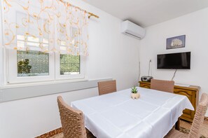 Un coin repas avec une table blanche, des chaises en osier, une petite plante en pot et un téléviseur fixé au mur.