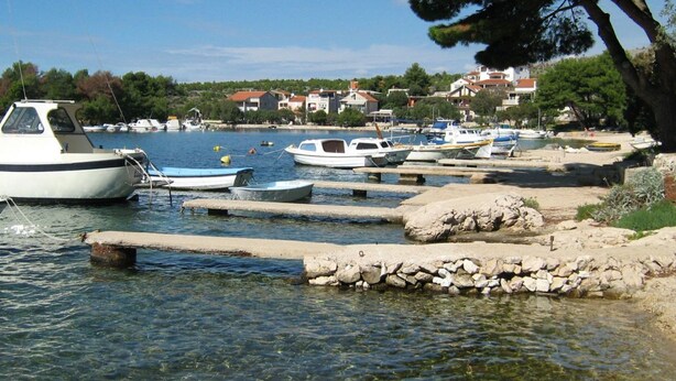 Un port de plaisance où sont amarrés plusieurs bateaux, avec une eau claire et une côte rocheuse, des maisons se dressant à l’arrière-plan.