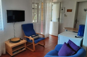 A living room with a television, a blue armchair, a small table with a bowl and remote, a blue sofa with purple cushions, and a white cabinet with a blue container. - Apartments Mala Kuca Kraj Mora (Sibenik)