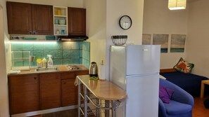 A compact kitchen with wooden cabinets, a stainless steel sink, and a white refrigerator. A small bar counter with a stool is in the center. A clock and artwork adorn the walls. - Apartments Mala Kuca Kraj Mora (Sibenik)