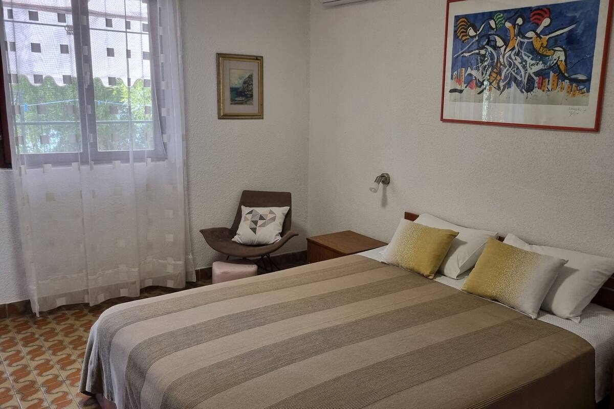 A hotel room with a bed, a chair, a nightstand, a wall-mounted lamp, and a painting.