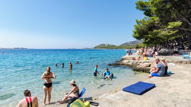 Ein Strand mit klarem Wasser, Menschen beim Schwimmen und Sonnenbaden, und eine felsige Küstenlinie mit Bäumen.