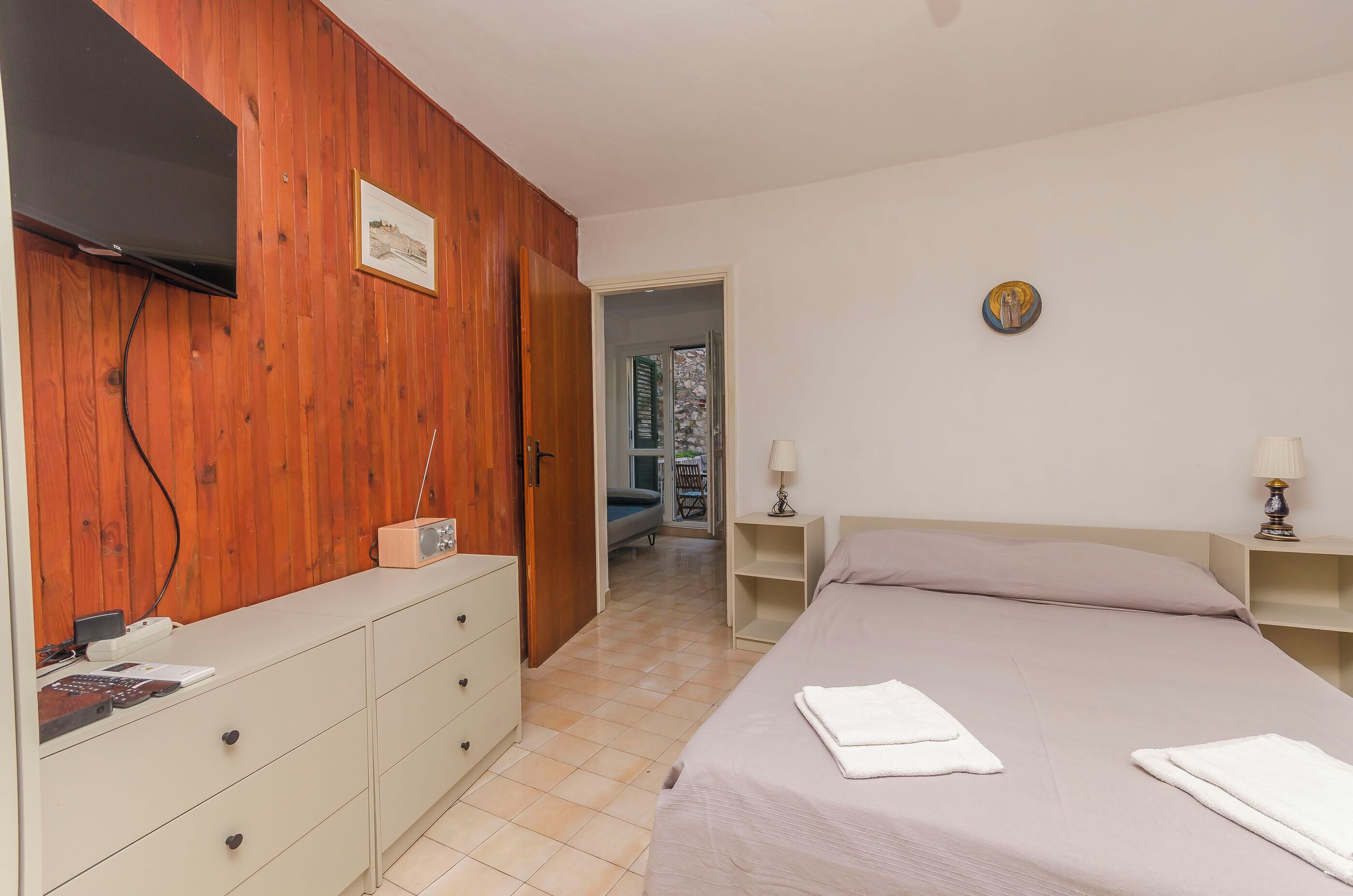 A bedroom with a bed, a dresser, a TV mounted on the wall, and a door leading to another room.
