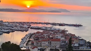 Eine Küstenstadt mit einem Yachthafen, Gebäuden mit roten Dächern und einem Sonnenuntergang über dem Wasser.