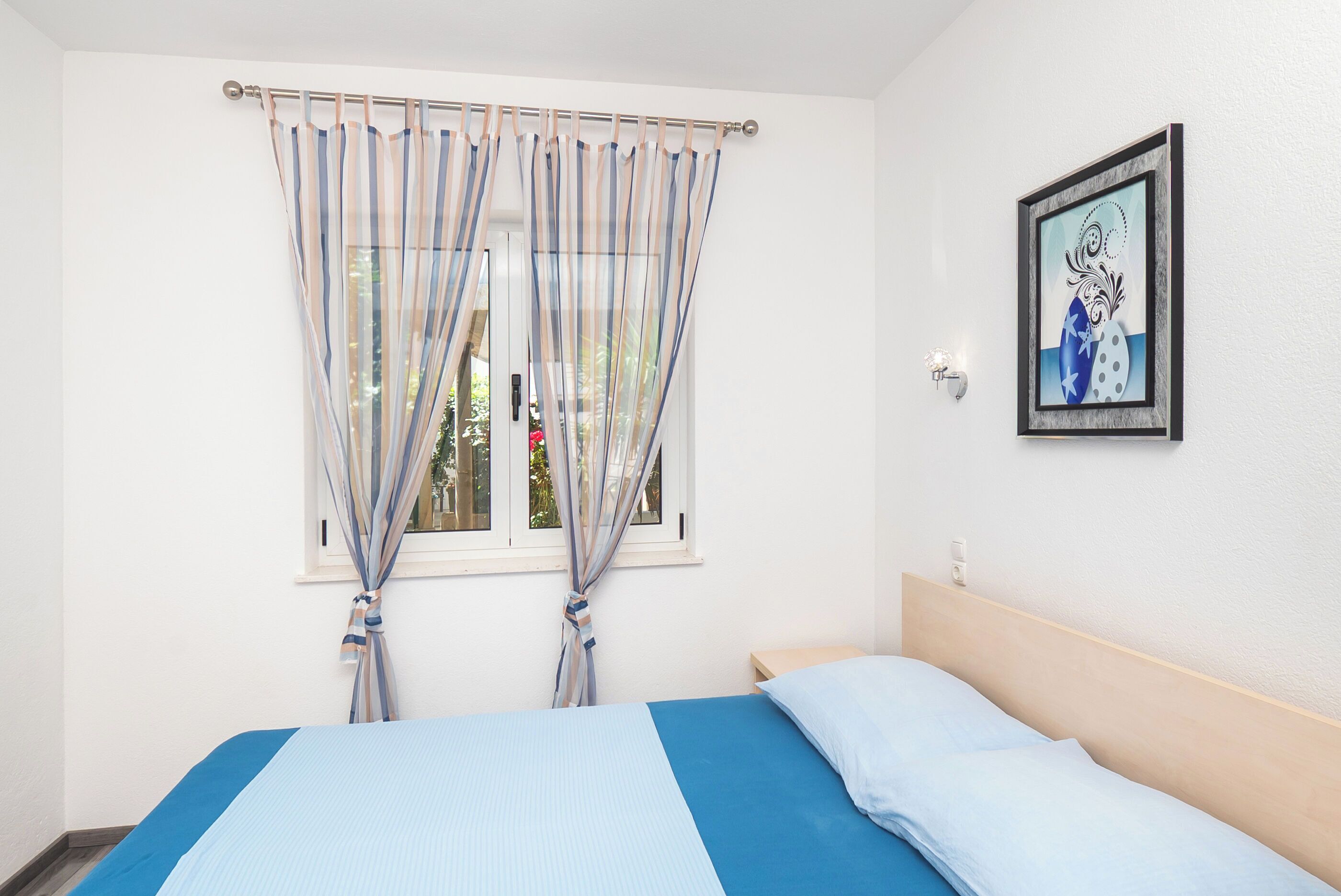 Een slaapkamer met een bed, gordijnen, een raam en een lijst met een foto aan de muur.