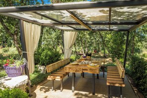 A covered outdoor dining area with a table and benches, surrounded by greenery. - Apartments Bijou (Umag)
