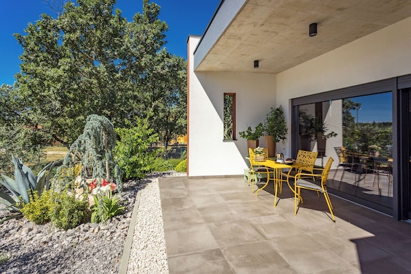 Une terrasse extérieure moderne, dotée d’un espace repas et entourée d’un jardin où poussent différentes plantes, ainsi que d’un grand arbre.