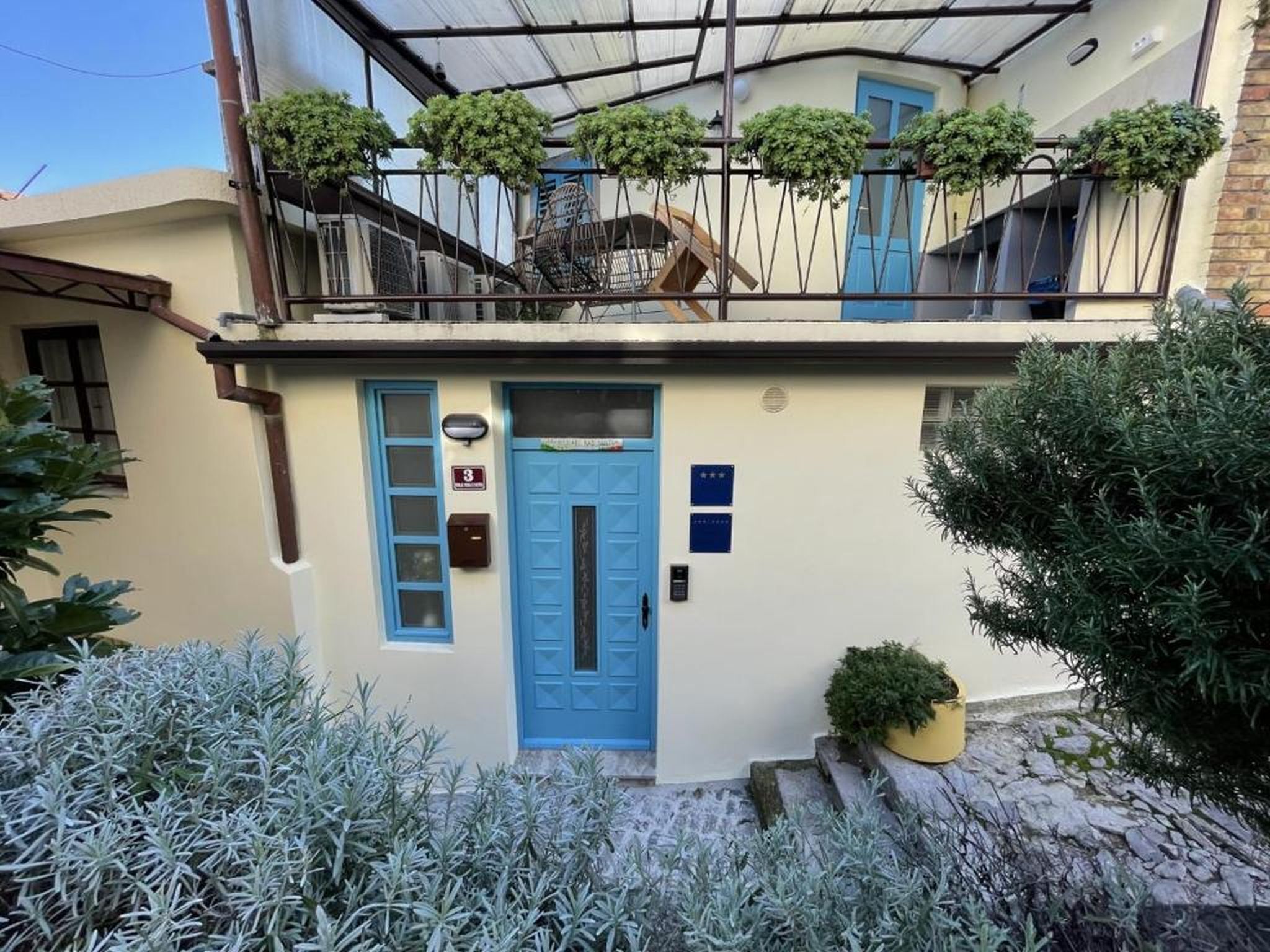 Ein zweistöckiges Gebäude mit einer blauen Tür, einem Balkon mit Blumentöpfen und einem kleinen Garten mit Sträuchern.