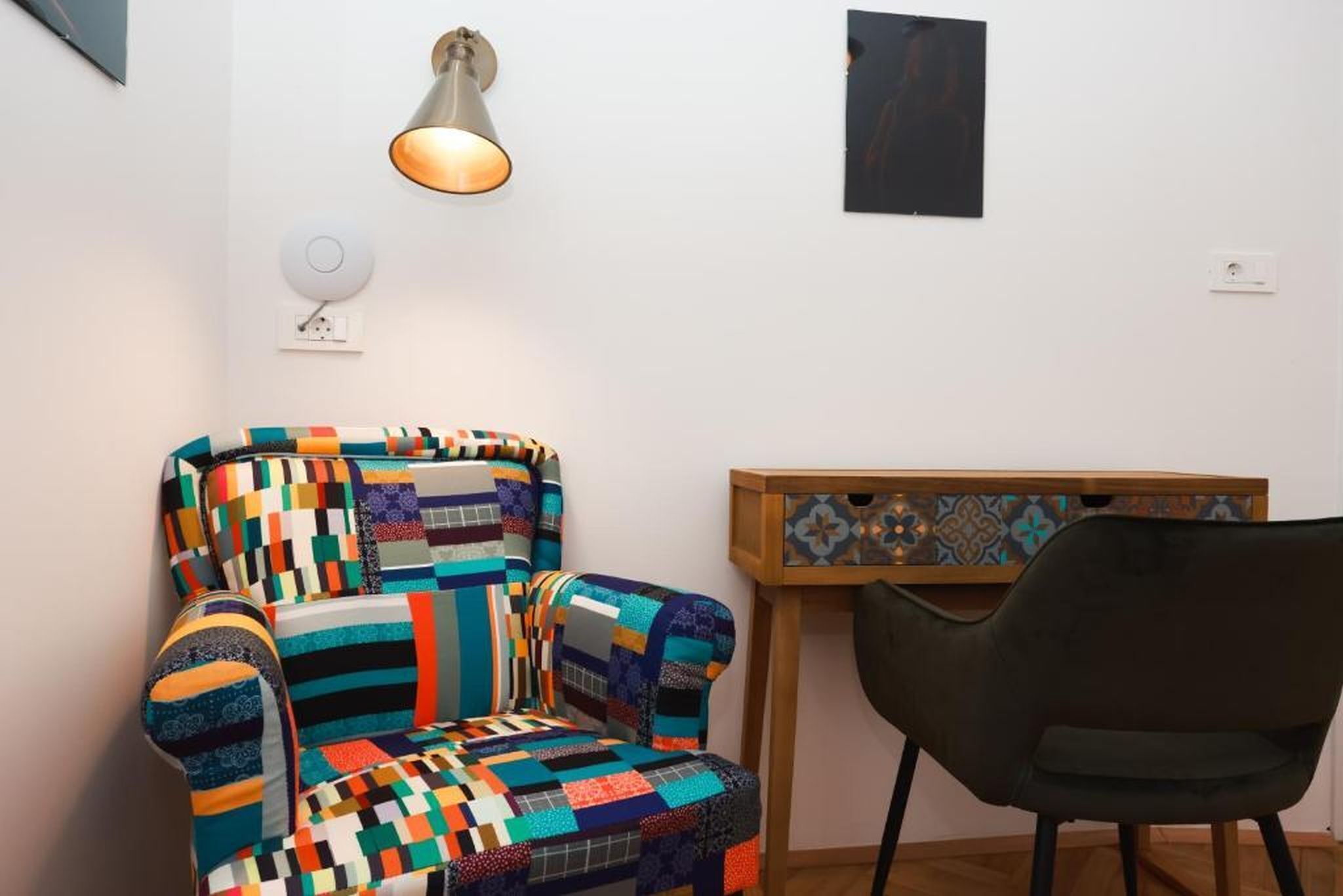 A room with a colorful patchwork chair, a black chair, a wooden side table with a patterned tray, and a wall-mounted lamp.