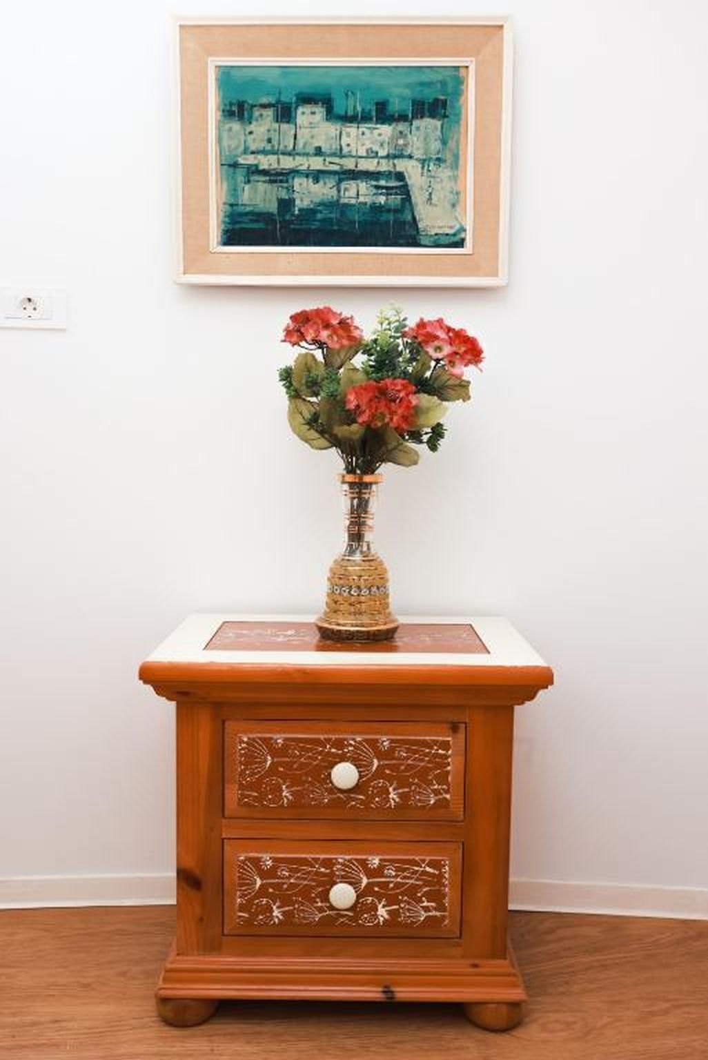 A wooden nightstand with a vase of red flowers, a framed painting on the wall, and a power outlet.