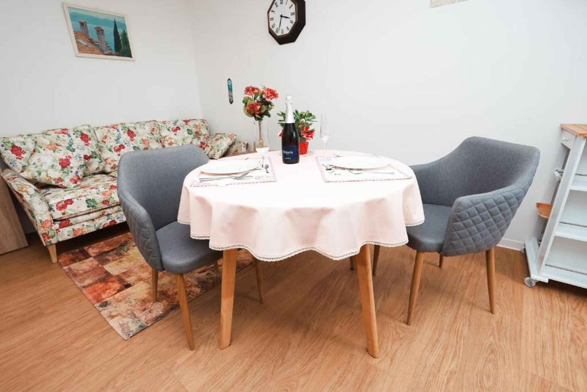 Un rincón de comedor con una mesa redonda, dos sillas, un sofá floral, un reloj y una pequeña planta en maceta.