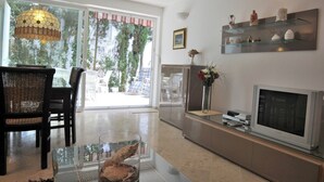 A modern living room with a glass coffee table, a flat-screen TV, wicker chairs, and a large window with a view of greenery. - Apartments Adrian (Rovinj)