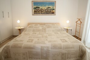 A bedroom with a patterned bedspread, two bedside lamps, a painting on the wall, and a wooden hanger on the right. - Apartments Adrian (Rovinj)