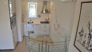 A modern kitchen with white cabinetry, a glass dining table, and a window providing natural light. - Apartments Adrian (Rovinj)