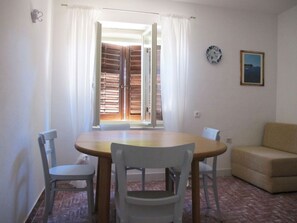 A dining area with a wooden table and white chairs, a beige sofa, and a view of a wooden door and window. - Apartments Marinic (Novalja)