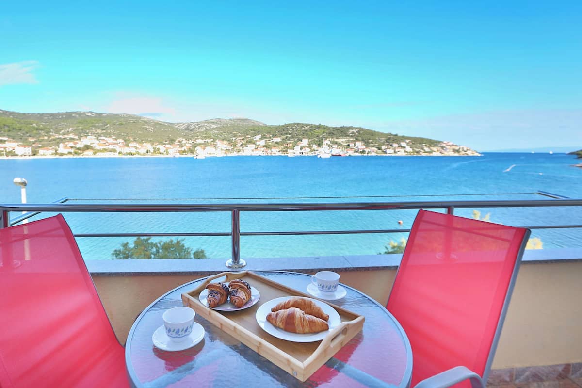 A balcony with a table set for breakfast, overlooking a coastal town and the sea.