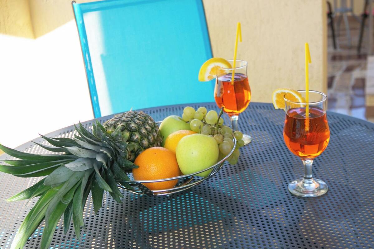 A table with a fruit bowl containing pineapple, oranges, and grapes, and two glasses of amber-colored drink with lemon slices and straws.