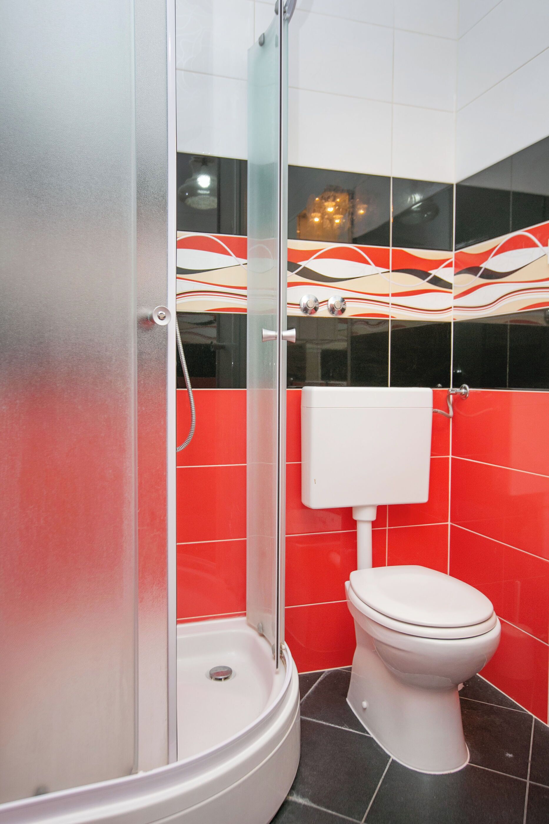 Um banheiro com chuveiro em gabinete de vidro, vaso sanitário branco e paredes com azulejos vermelhos e pretos.