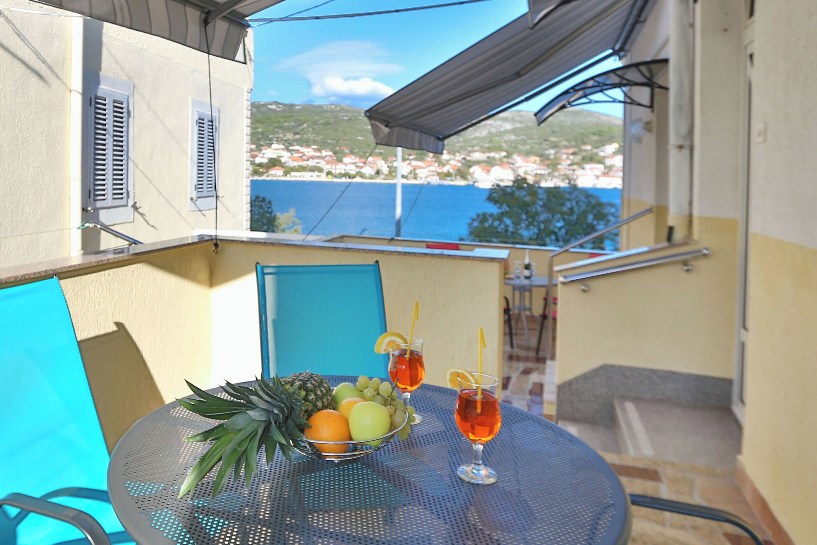 A balcony with a table set with fruit and drinks, overlooking a coastal town.