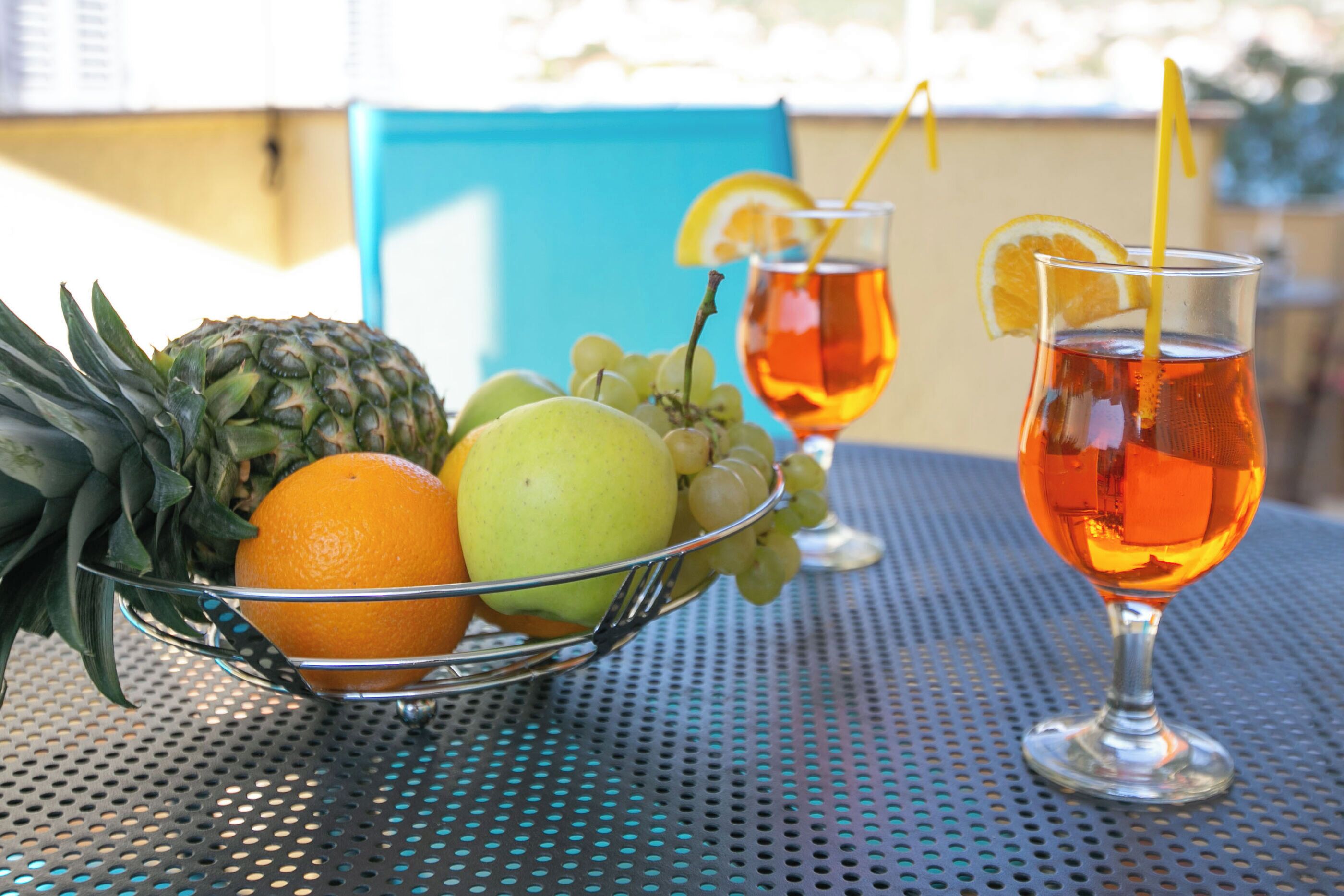 Ein Tisch mit einer Obstschale, die eine Ananas, eine Orange und einen grünen Apfel enthält, sowie zwei Teegläser mit Zitronenscheiben und Halmsaugrohren.