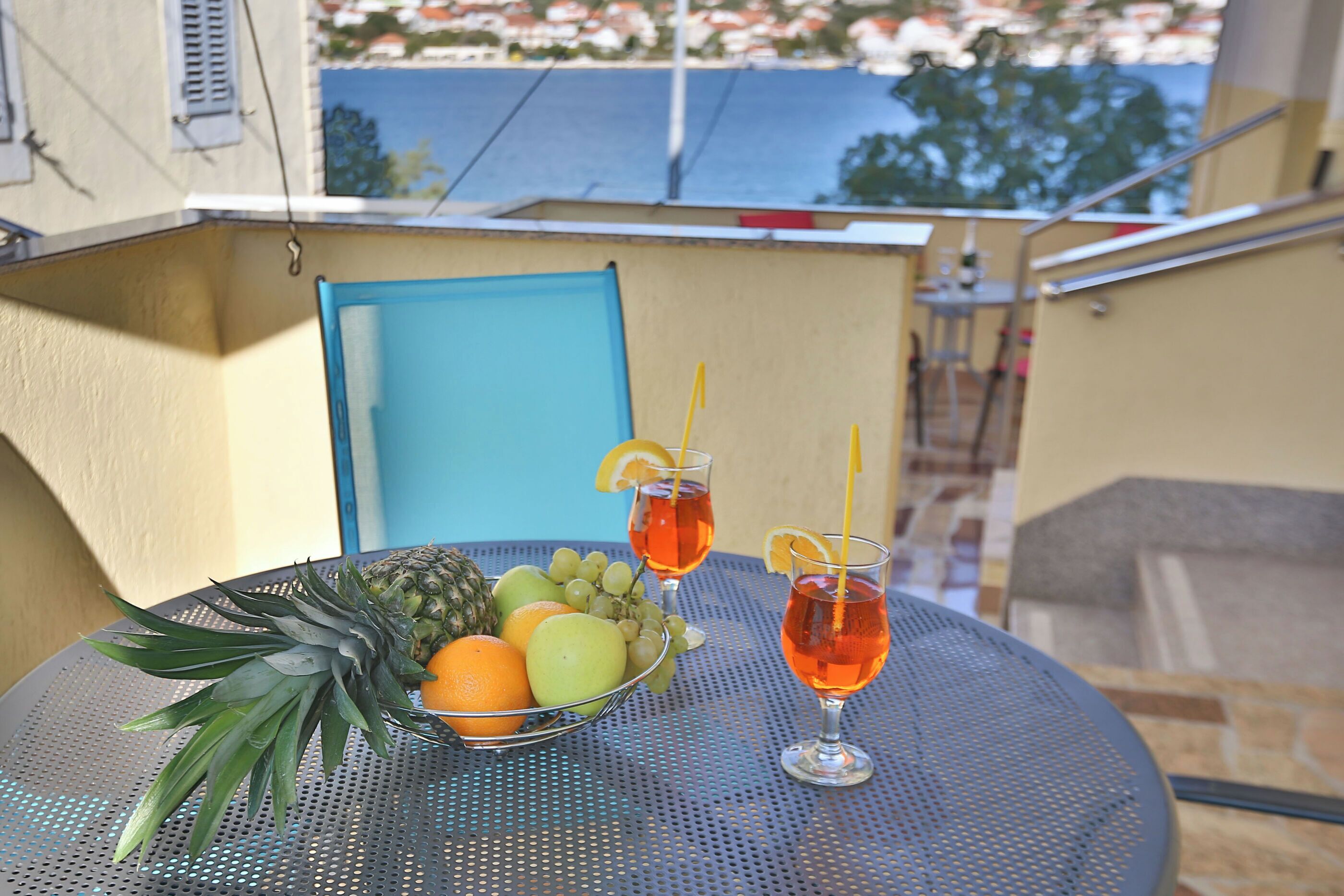 Une table ronde en métal sur laquelle se trouve un saladier rempli d’ananas, d’oranges et de raisins, ainsi que deux verres contenant une boisson rouge, accompagnés de pailles et de rondelles de citron.