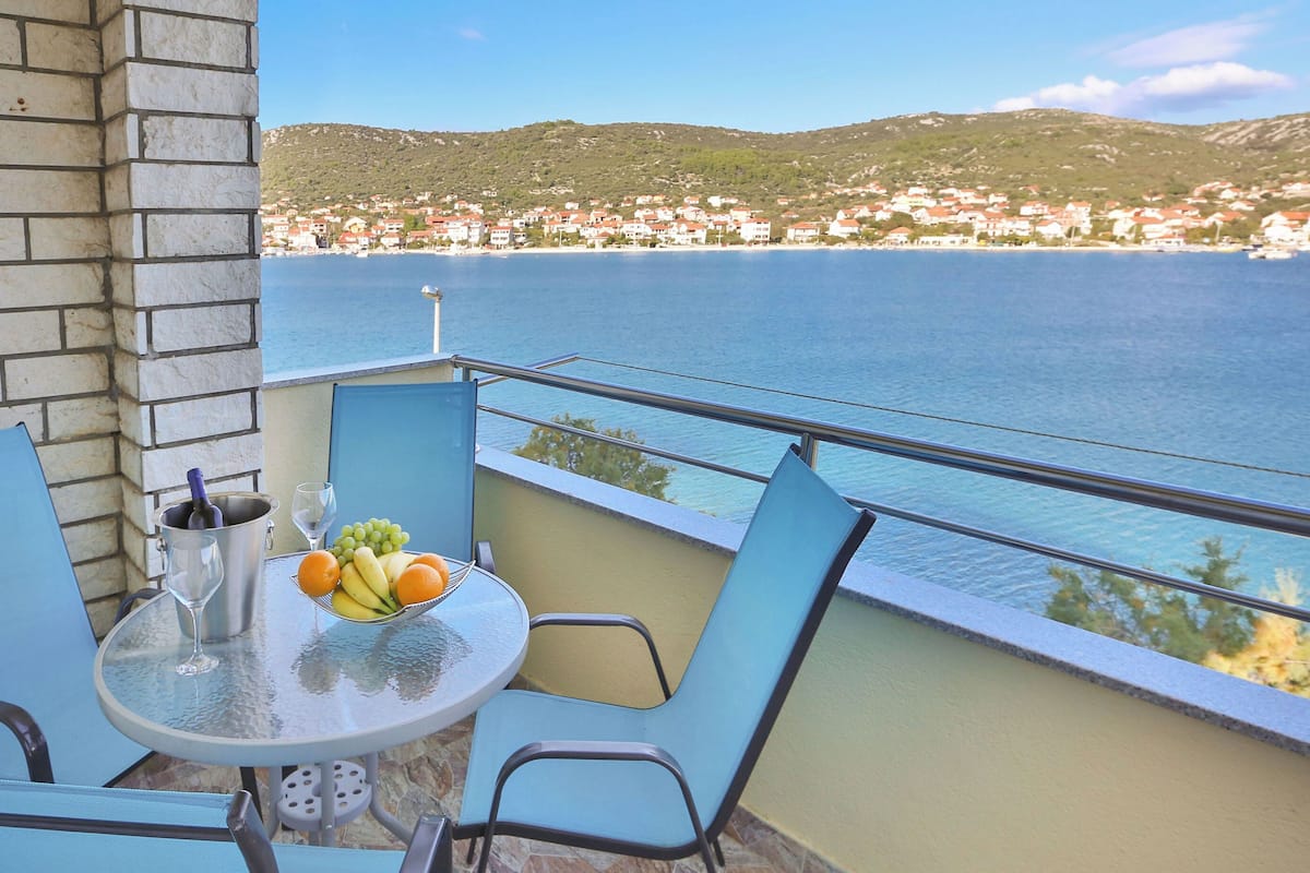 A balcony with a table set for two, a bottle of wine, and a bowl of fruit. The view includes a coastal town and a body of water.