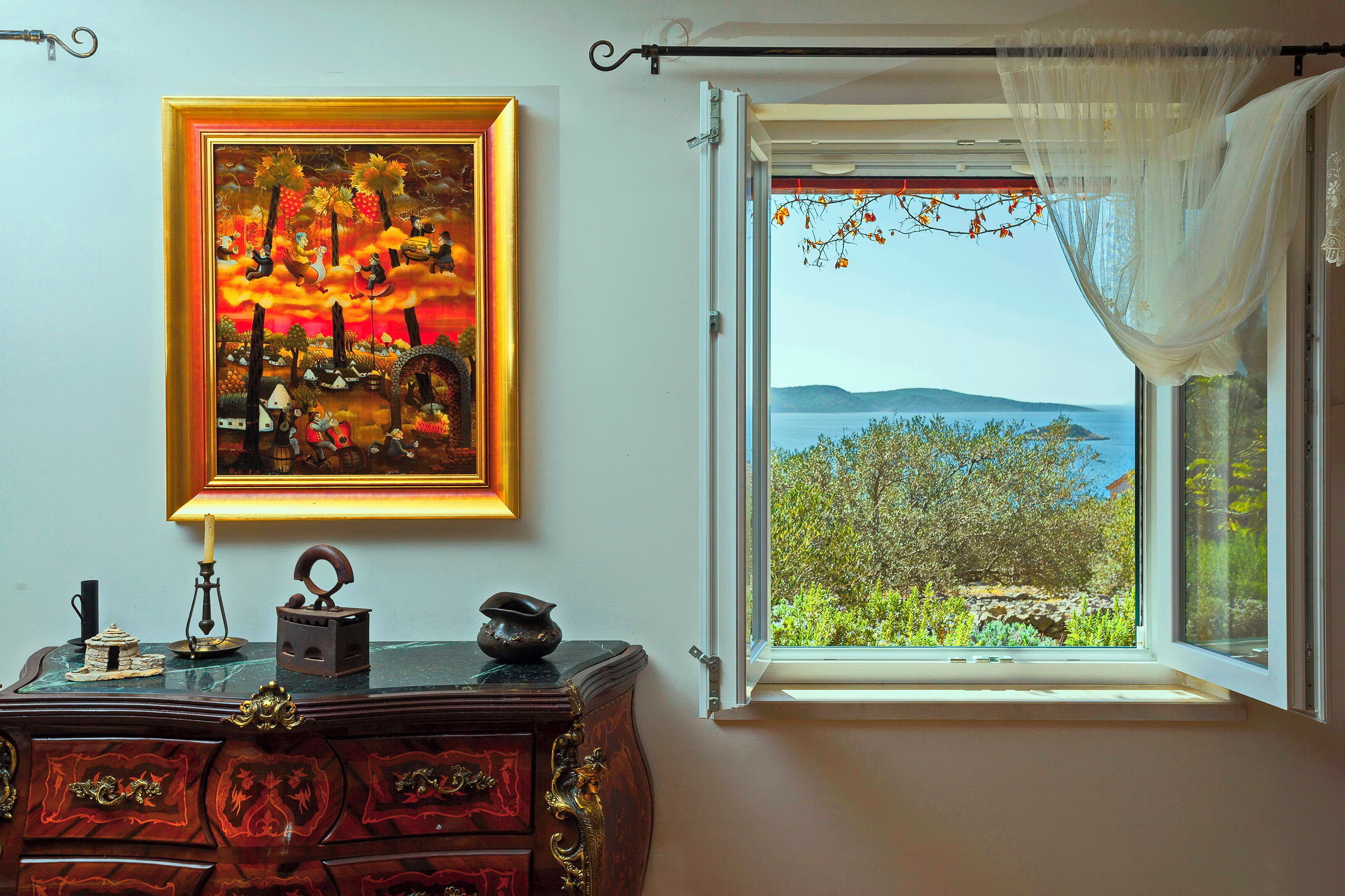 A room with a view of a landscape through a window, a painting on the wall, and a wooden dresser with drawers.