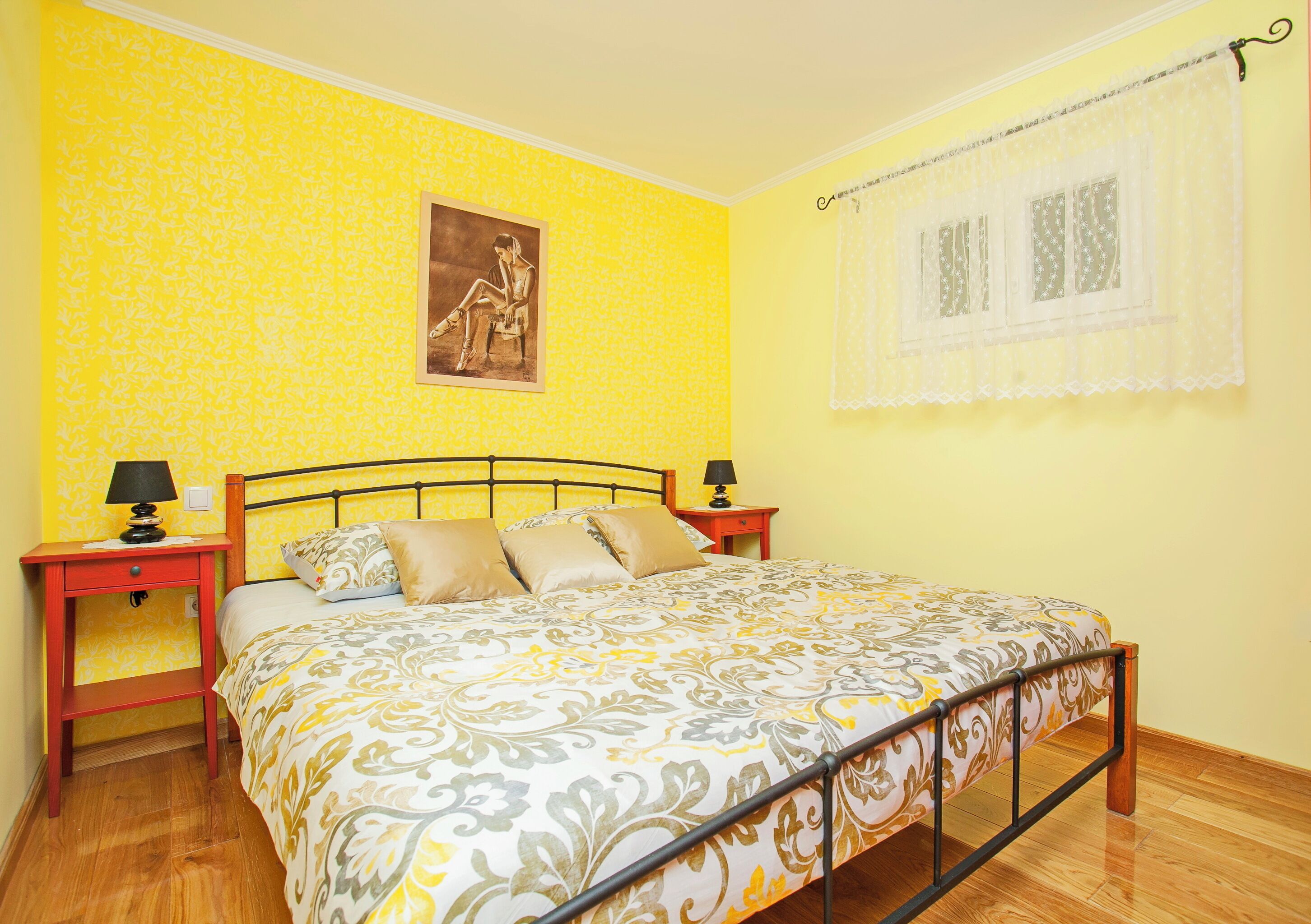 A bedroom with a bed, two bedside tables, a nightstand, a window with lace curtains, and a wall with a patterned wallpaper.