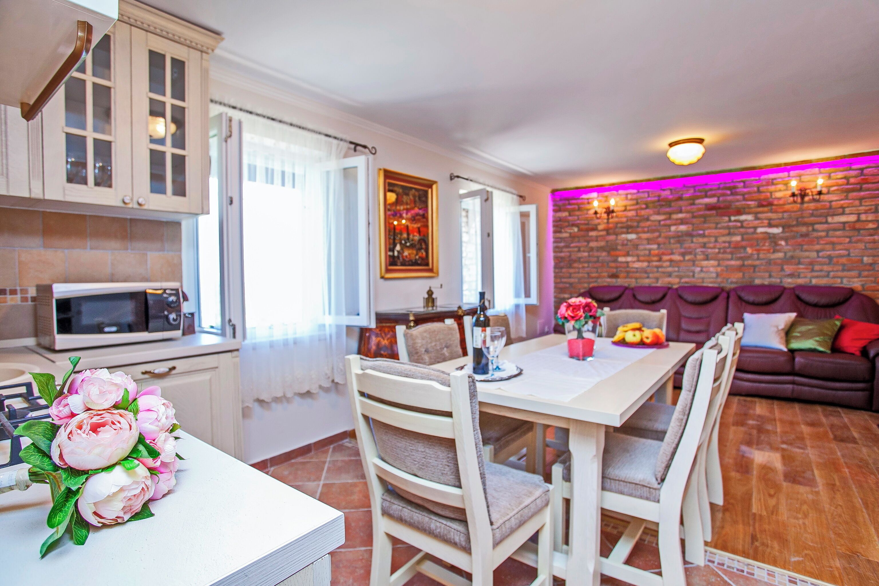 A modern kitchen with white cabinets, a dining table with chairs, and a brick wall with a neon light.