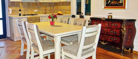 A dining area with a wooden table and white chairs, a kitchen with white cabinets, and a painting on the wall.