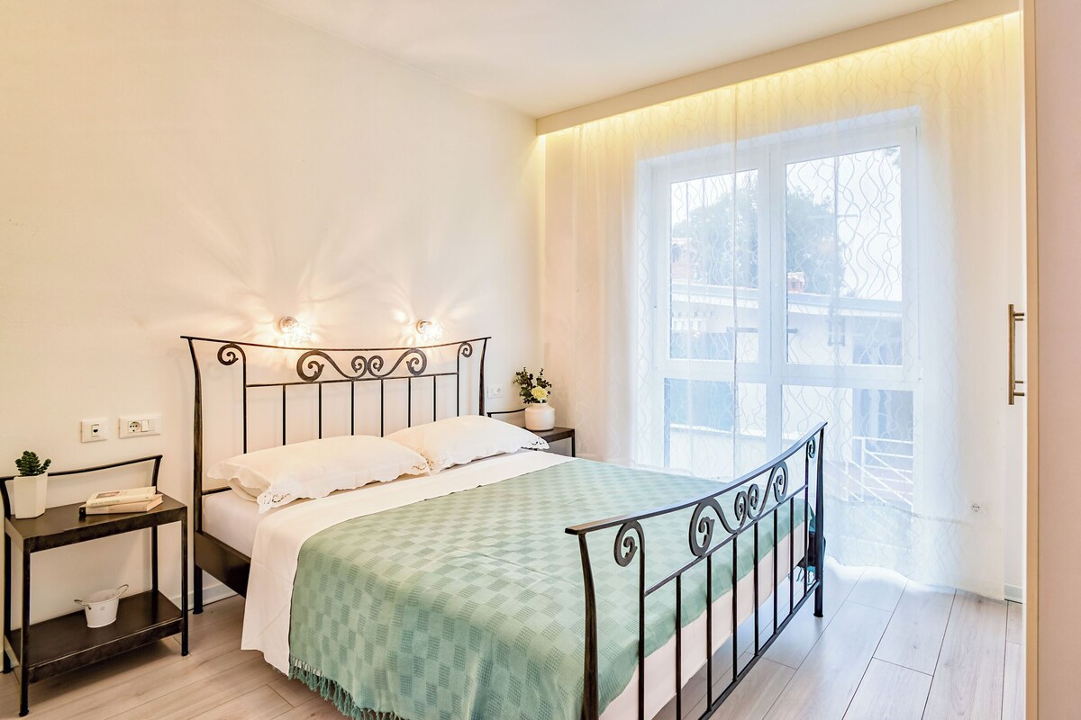 A bedroom with a bed, bedside tables, a window with curtains, and a view of the outside.