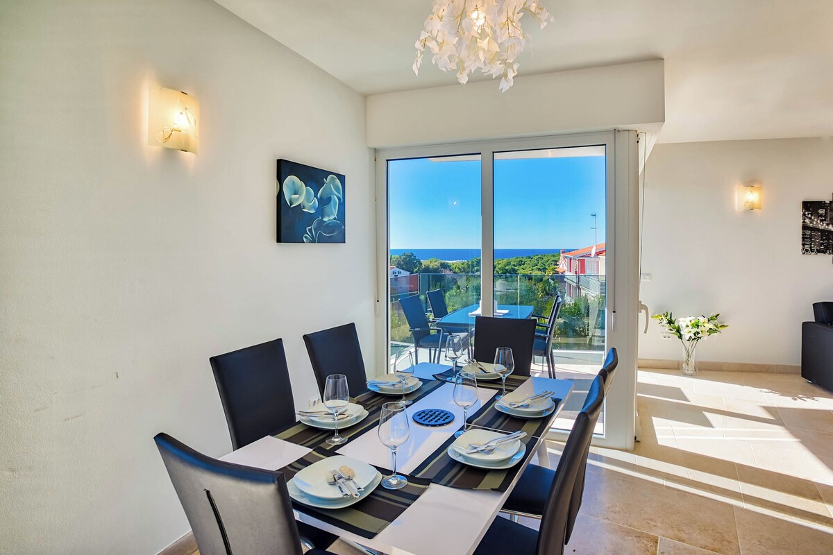A dining area with a table set for six, a view of the outdoors, and a chandelier.
