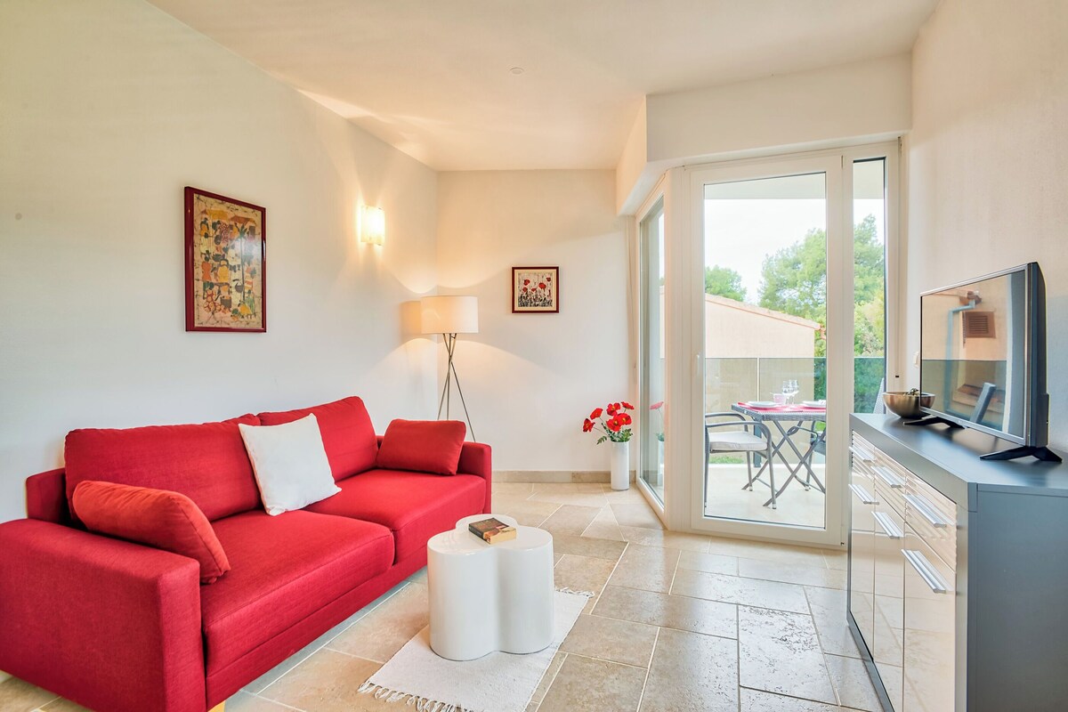 A modern living room with a red sofa, a small white side table, a TV, and a glass door leading to an outdoor patio.
